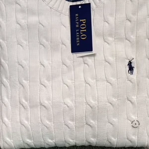 Vit kabelstickad tröja Polo Ralph Lauren - Snygg vit kabelstickad tröja från Polo Ralph Lauren med klassisk blå logga på bröstet. Tröjan har rund hals, ribbade muddar och är långärmad. Perfekt för dig som gillar stilrena och tidlösa plagg med premiumkänsla.