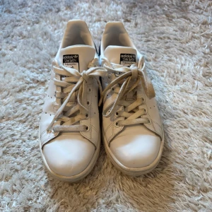 Adidas Stan Smith vita sneakers - Klassiska Adidas Stan Smith sneakers i vitt skinn med svarta detaljer på hälen och perforerade ränder på sidorna. Skorna har snörning och en platt sula, perfekt för en clean och tidlös look. Ikonisk logga på plösen och hälen.