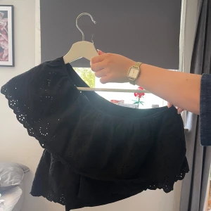 Svart volangig offshoulder blus - Supersöt svart blus med volanger och broderade detaljer. Offshoulder-modell med luftig passform och vackert hålmönster i tyget. Perfekt för dig som gillar romantisk och trendig stil. T-shirten är från Mango