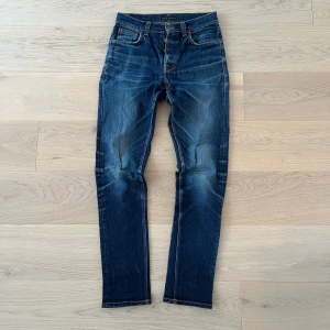 Nudie Jeans Steady Eddie blå jeans - Säljer ett par Nudie Jeans Steady Eddie i klassisk blå tvätt med snygga slitningar och orange sömmar. Jeansen har rak passform, fem fickor och är tillverkade i 99% bomull och 1% elastan för lite stretch. Perfekta för dig som gillar en tidlös jeanslook. Hål i högerfickan, men inget som hindrar från att ha saker i den. 