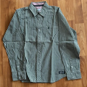 Barn - Rutig skjorta från Jonas Nielsen M - Barn - Grön och vit rutig skjorta från Jonas Nielsen Stockholm i bomull. Klassisk krage med kontrastfoder i rött och vitt, bröstficka med broderad detalj och tryck på ryggen med lejon och texten J.N.S.B College Team. Långärmad och knäppning framtill.