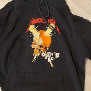 Svart Metallica hoodie Damage Inc. - Svart hoodie med Metallica Damage Inc.-tryck fram och turnédatum bak. Stor färgglad dödskalle med orange och gult motiv på bröstet, röda Metallica-bokstäver och snörning i huvan. Perfekt för dig som älskar bandmerch och vill sticka ut.