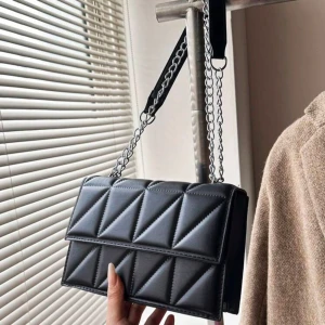 Svart quiltad axelväska med kedja - Stilren svart axelväska med quiltad design och coola triangeldetaljer. Väskan har en silverfärgad kedjerem och bred axelrem i tyg för extra komfort. Perfekt för att lyfta vilken outfit som helst.