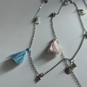 Silverfärgat halsband med tofsar och berlocker - Unikt halsband i silverfärgad metall med små berlocker, stjärnor och runda plattor. Dekorerat med två tofsar i ljusblått och ljusrosa samt pärlliknande detaljer i vitt och blått. Perfekt för dig som gillar att sticka ut med coola accessoarer.