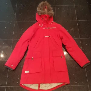 Röd parkas från Didriksons med fuskpäls - Riktigt snygg röd parkas från Didriksons med huva och fuskpäls, perfekt för kalla dagar. Jackan har stora fickor, dragkedja och snörning i midjan. Insidan är quiltad och beige, vilket ger extra värme och stil. Klassisk look med praktiska detaljer. I fickan som har en vit dragkedja på andra bilden finns det ett hål, men som sagt är det i fickan så det kommer inte synas. Nypris 1500kr. PRIS KAN DISKUTERAS!