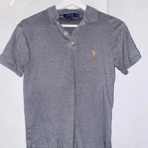 Grå Polo Ralph Lauren t-shirt - Snygg grå t-shirt från Polo Ralph Lauren i slim fit med klassisk krage och korta ärmar. T-shirten har knappar vid halsen och den ikoniska broderade loggan på bröstet. Tillverkad i mjuk bomull som är skön mot huden.