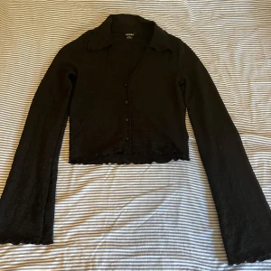 Svart croppad kofta från Monki - Svart tröja från Monki i croppad modell med långärmade, lätt utsvängda ärmar och vågig kant vid ärmslut och nederkant. Tröjan har knappar framtill och är gjord i ett tunt, strukturerat syntetmaterial. Aldrig använd.