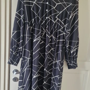 Svartvit långklänning från SHEIN XL - Svart långklänning från SHEIN i storlek XL med vitt grafiskt mönster. Klänningen har långa ärmar med smock vid ärmslut, knappar framtill och en avslappnad passform. Perfekt för dig som gillar statement-plagg med cool design.