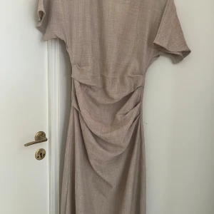 Beige långklänning med drapering - Snygg beige långklänning med korta ärmar och draperad midja som ger en cool siluett. Klänningen har en stilren look med mjukt, vävt material och passar perfekt för dig som gillar minimalistisk stil. Enkel att styla med accessoarer för en trendig vibe. Helt ny, oanvänt.