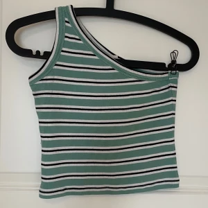 Randig one shoulder topp i grönt - Snygg ribbad one shoulder topp med gröna, vita och svarta ränder. Modellen är croppad och har en axelbandslös design på ena sidan. Perfekt för dig som gillar en trendig och unik look. Materialet är mjukt och stretchigt för en bekväm passform.