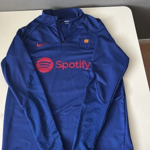 Blå långärmad FC Barcelona Nike tröja - Säljer en blå långärmad FC Barcelona träningströja från Nike med Dri-FIT material. Tröjan har halv dragkedja, klubbmärke på bröstet och röd Nike-logga samt Spotify-tryck framtill. Slim fit-modell, perfekt för dig som älskar Barca.