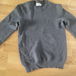 Grå ribbstickad tröja från East West - Säljer en stilren grå ribbstickad tröja från East West. Tröjan har rund halsringning, lång ärm och en clean look med diskreta horisontella ränder i stickningen. Perfekt för dig som gillar enkel och snygg stil.