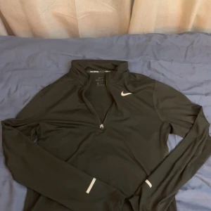 Svart Nike half zip träningströja - Svart långärmad träningströja från Nike med half zip och Dri-FIT-material. Snygg vit Nike-logga på bröstet och reflekterande detaljer på ärmarna. Perfekt för löpning eller gymmet, sitter skönt och andas bra.