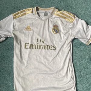 Real Madrid Hazard 7 Adidas tröja - Säljer en officiell Real Madrid matchtröja från Adidas med Hazard 7 på ryggen. Tröjan är vit med guldiga detaljer, klubbmärke och Adidas-logga på bröstet samt Fly Emirates-tryck. Klassisk passform och korta ärmar, tillverkad i lätt och ventilerande material.