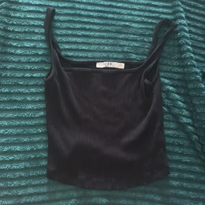 Svart ribbad linnetopp från NA-KD - Svart ribbad linnetopp från NA-KD med smala axelband och croppad passform. Toppen är enkel men stilren, perfekt att matcha med jeans eller kjol. Materialet är stretchigt och följsamt för en bekväm känsla.