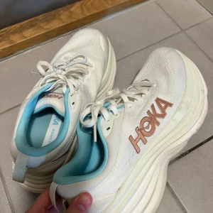 Vita Hoka One One Bondi 8 sneakers - Snygga Hoka One One Bondi 8 sneakers i vitt med ljusblå insida och detaljer. Skorna har en chunky, extra tjock sula och HOKA-logga i kopparfärg på sidan. Perfekta för dig som gillar komfort och stil när du är ute och rör på dig.