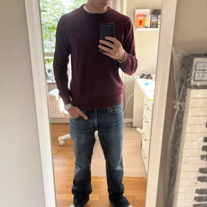 Vinröd stickad tröja från GANT - Snygg vinröd stickad tröja från GANT med rund hals och diskret logga broderad på bröstet. Tröjan har långärmade ärmar och ribbade muddar vid hals, ärmslut och nederkant. Perfekt för dig som gillar klassisk stil med en modern touch.