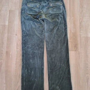 Gröna velourbyxor Juicy Couture S - Mysiga gröna velourbyxor från Juicy Couture med raka ben och stora fickor både fram och bak. Byxorna har broderad JC-logga på ena fickan och coola detaljer med knappar på bakfickorna. Perfekta för en chill och trendig look.