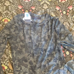 Camo långärmad tröja från Primark - Snygg långärmad tröja från Primark i grått och blått camouflagemönster. Tröjan har en klassisk rund halsringning och är tillverkad i mjuk bomull, perfekt för en chill och avslappnad stil.