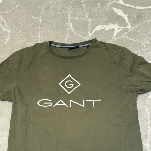 Olivgrön GANT t-shirt med logga - Snygg olivgrön t-shirt från GANT med stor vit logga framtill. Klassisk rund halsringning och korta ärmar. Skön och mjuk känsla, perfekt till jeans eller shorts. Enkel men stilren design som funkar året runt.
