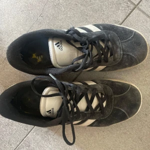 Svarta och vita Adidas sneakers - Klassiska Adidas sneakers i svart mocka med vita detaljer och de ikoniska tre ränderna på sidorna. Skorna har svart snörning, vit platt sula och rund tå. 