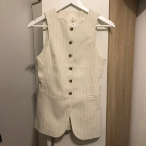 Randig beige väst från H&M - Snygg beige väst från H&M med tunna vertikala ränder och bruna knappar framtill. Västen har två fickor och en figursydd, något längre passform. 