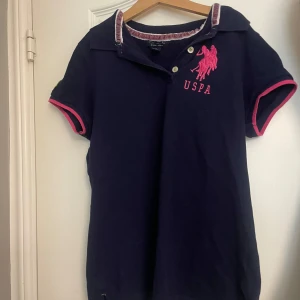 Marinblå pikétröja från USPA - Snygg marinblå pikétröja från USPA med rosa detaljer och broderad logga på bröstet. Krage med knappar, korta ärmar med rosa kant och en grön broderad siffra 3 på ena ärmen. Perfekt för en sportig och stilren look. Så himla snyggt till ett par lågmidjade jeans. Storleken är i S. Varan säljs på flera plattformar💕