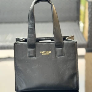 NY Svart handväska  - Helt ny stilren svart handväska i skinnimitation. Väskan har raka linjer, dubbla handtag och ett lila innerfoder med dragkedjefack. Finns även axelrem i guldkedja som man kan ta av/på. Ej på bild tyvärr. Se bilder med linjal för storlek/mått. Hämtas i Hägersten. 