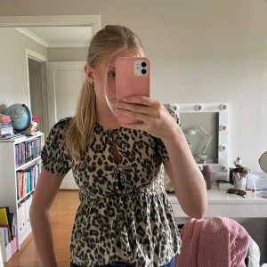 Leopardmönstrad blus med knyt - Säljer en beige och svart leopardmönstrad blus med kort puffärm och två knytningar framtill. Modellen har rund halsringning och lätt volang nedtill för en söt och trendig look. Perfekt för dig som vill sticka ut lite extra.