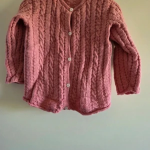 Rosa stickad kofta med knappar - Mysig rosa stickad kofta med kabelstickat mönster och vita knappar framtill. Koftan har långärmade ärmar och en klassisk rund halsringning. Perfekt för att hålla sig varm och ändå se chill ut. Passar dig som gillar en soft och feminin stil.