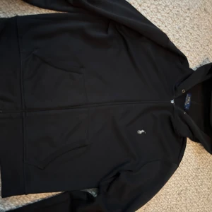 Svart hoodie från Polo Ralph Lauren - Svart hoodiejacka från Polo Ralph Lauren med dragkedja framtill och två fickor. Klassisk logga broderad på bröstet. Perfekt för chill dagar och enkel att matcha med jeans eller joggers. Passar även storlek M