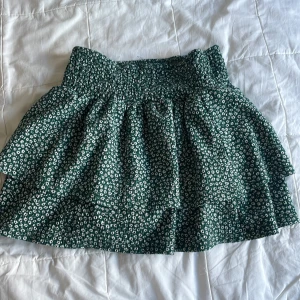 Grön blommig kjol från Zara - Supersöt grön kortkjol från Zara med vitt blommigt mönster. Kjolen har smockad midja och volangkant som ger en skön och flowy känsla. Perfekt för dig som gillar en lekfull och fräsch stil. Materialet är mjukt och lätt, passar perfekt till varma dagar.