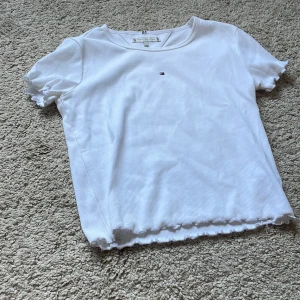 Vit ribbad topp Tommy Hilfiger - Supersöt vit ribbad topp från Tommy Hilfiger med korta ärmar och volangkant både vid ärmslut och nederkant. Liten broderad logga framtill. Perfekt till jeans eller kjol för en clean och fräsch look.