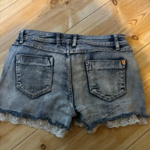 Grå jeansshorts med spetskant - Snygga grå jeansshorts med slitna detaljer och rå kant, dekorerade med vit spets längst ner. Klassisk femficksmodell med dragkedja och knapp framtill. Perfekt för dig som vill ha en cool och feminin stil på sommaren.