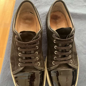 Svarta sneakers med lackad tå - Säljer ett par svarta sneakers med snyggt lackad tå och ovandel i mocka. Skorna har rund tå, snörning och vit platt sula som ger en clean kontrast. Insidan är fodrad i beige skinn och detaljerna är diskreta men stilrena. Perfekt för dig som gillar en unik touch på klassiska sneakers.