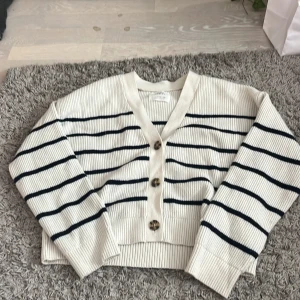 Vit och svart randig kofta med knappar - Säljer en ribbad kofta från Zara  i vit med svarta horisontella ränder. Koftan har v-ringning, tre stora knappar framtill och långa ärmar. Materialet är mjuk stickad bomull, perfekt för lager-på-lager och chill dagar.