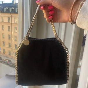 Svart Stella McCartney väska - Säljer en svart axelväska från Stella McCartney med guldig kedjaoch snygga detaljer längs kanterna. Sparsamt använd strl tiny! Nypris runt 10000
