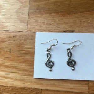 Silvriga örhängen med musikmotiv - Ett par coola örhängen i silverfärgad metall med hängande G-klav som motiv. Perfekt för dig som älskar musik och vill ha något unikt till din stil. Krokarna är enkla att sätta i och passar till alla tillfällen. Snygg detalj som sticker ut!Jag själv är nickelallergiker och har inte haft några problem. 