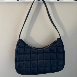 Svart axelväska  - Stilren svart axelväska med quiltad design och silvrig dragkedja. Väskan har en mjuk, fyrkantigt vadderad yta och ett rymligt innerfack med extra ficka. Perfekt för att lyfta din outfit med en trendig touch. Knappast använd 