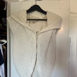 Vit stickad väst med huva - Mysig vit stickad väst/kofta. Koftan har olika stickade strukturer och är ärmlös, perfekt att slänga över en långärmad tröja för extra värme. Snygg och enkel att styla till många outfits. Aldrig använd endast testad 