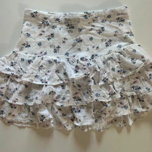 Vit blommig volangkjol  - Supersöt kort kjol i vit bomull med små blå och rosa blommor. Kjolen har flera lager volanger och en smockad midja som ger en skön och flexibel passform. Perfekt för dig som gillar romantisk och feminin stil.