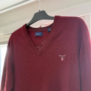 Vinröd stickad tröja från GANT - Snygg vinröd stickad tröja från GANT med v-ringad hals och broderad logga på bröstet. Tröjan har långa ärmar och ribbade muddar vid hals, ärmslut och nederkant. Perfekt för dig som gillar klassisk stil med en modern touch.