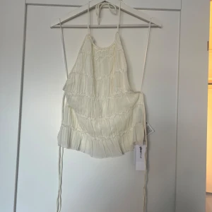 Vit linnetopp från Gina Tricot - Supersöt vit linnetopp från Gina Tricot med tunna axelband och volangiga lager. Toppen har knytband i ryggen och en lätt, flowy känsla. Perfekt för varma dagar och ger en romantisk vibe till din outfit.
