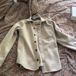 Beige overshirt med ficka - Snygg beige overshirt med klassisk krage och svarta knappar framtill. Skjortan har en bröstficka och är tillverkad i ett mjukt material som ger en chill och stilren vibe. Perfekt att slänga över en t-shirt för en avslappnad look.
