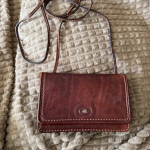 Brun axelväska i äkta skinn från The Bridge - Säljer en klassisk brun axelväska från The Bridge i äkta skinn. Väskan har ett stilrent, rektangulärt format med lock och magnetknäppning, samt justerbar axelrem. Invändigt finns flera fack och dragkedja för smart organisering. Perfekt för dig som gillar tidlös och snygg design.