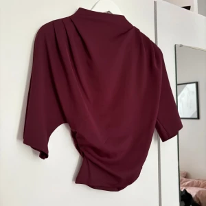Vinröd blus  - Säljer en vinröd blus med lös passform och trekvartsärmar. Toppen har en snygg drapering på ena sidan och hög halsringning. Perfekt för dig som gillar stilrena plagg med en twist. Materialet känns mjukt och följsamt mot huden.