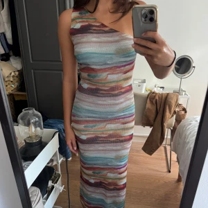 Randig långklänning från Zara - Säljer en snygg långklänning från Zara med färgglada, horisontella ränder i blått, grönt, vinrött, orange och beige. Klänningen har oneshoulder-design och är figurnära med stretchigt material. Perfekt för dig som gillar unika mönster och stilren passform.