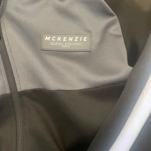 Svart och grå vindjacka från McKenzie - Snygg svart och grå vindjacka från McKenzie med huva och dragsko. Jackan har hel dragkedja framtill, ribbade muddar och en grå rand längs ärmarna. Perfekt för en sportig look och chill vardagsstil.