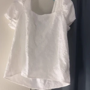 Vit blus med puffärm och svart rosett - Supersöt vit blus med puffiga korta ärmar och subtilt blommigt mönster i tyget. Blusen har en fyrkantig halsringning och en stor svart rosett bak som ger en extra cool touch. Materialet känns lyxigt och har en lätt struktur. Perfekt för dig som gillar statement-detaljer.