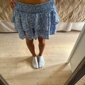 Blommig blå volangkjol  - Supersöt kort kjol från Kappahl i blått med vitt blommigt mönster. Kjolen har bred smock i midjan och flera lager med volanger som ger en flowy och lekfull look. Perfekt för varma dagar och enkel att matcha med en crop top eller t-shirt. Öppen för prisförslag